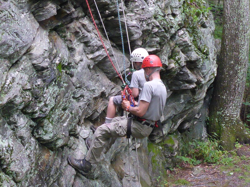 Two people are rappelling down a rocky cliff face. Both individuals are wearing helmets, one white and one red, and are secured with ropes and harnesses. The person in the foreground is wearing a grey t-shirt and khaki pants, while the other person is wearing a light-colored t-shirt. The cliff is textured with moss and lichen, and trees are visible in the background, suggesting an outdoor setting.